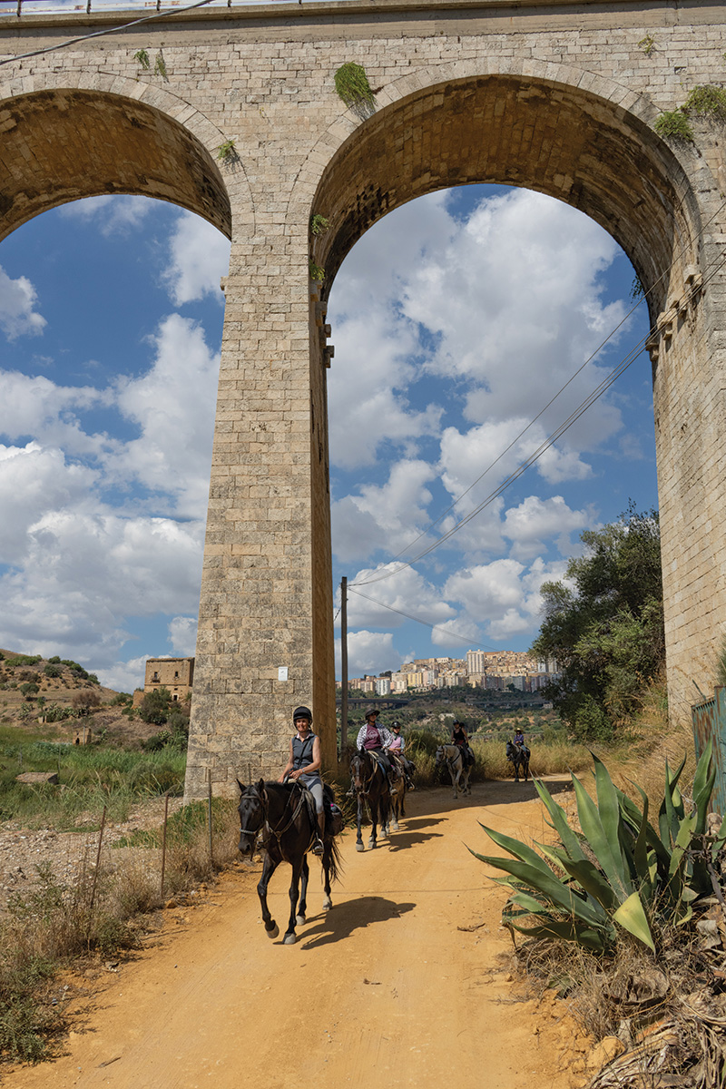 Horseback riders follow Sicily's Imera river valley under massive highway bridges