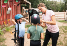 A worker at a riding stable helps young equestrians with their helmets.