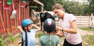 A worker at a riding stable helps young equestrians with their helmets.