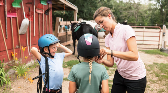 A worker at a riding stable helps young equestrians with their helmets.