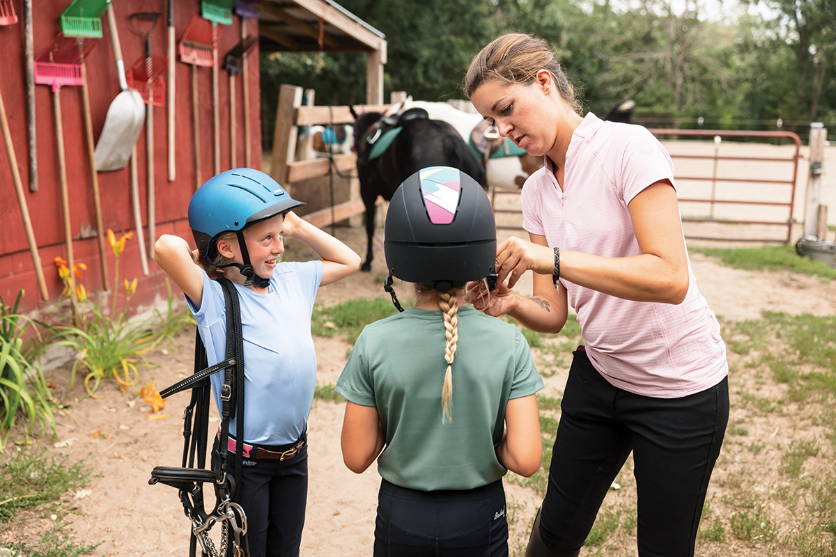 A worker at a riding stable helps young equestrians with their helmets.